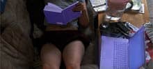 Frau liegt auf dem Sofa und liest ein Buch, neben dran ein Laptop (auf dem wahrscheinlich ihr Facebook-Account wartet)